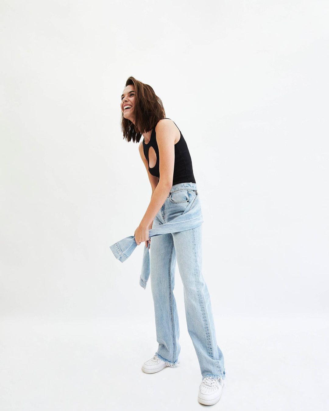 Smiling woman in a black tank top and light wash denim pants stands indoors in a brightly lit studio, looking up and to her left.