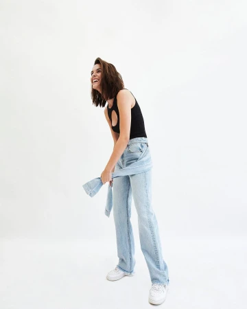 Smiling woman in a black tank top and light wash denim pants stands indoors in a brightly lit studio, looking up and to her left.