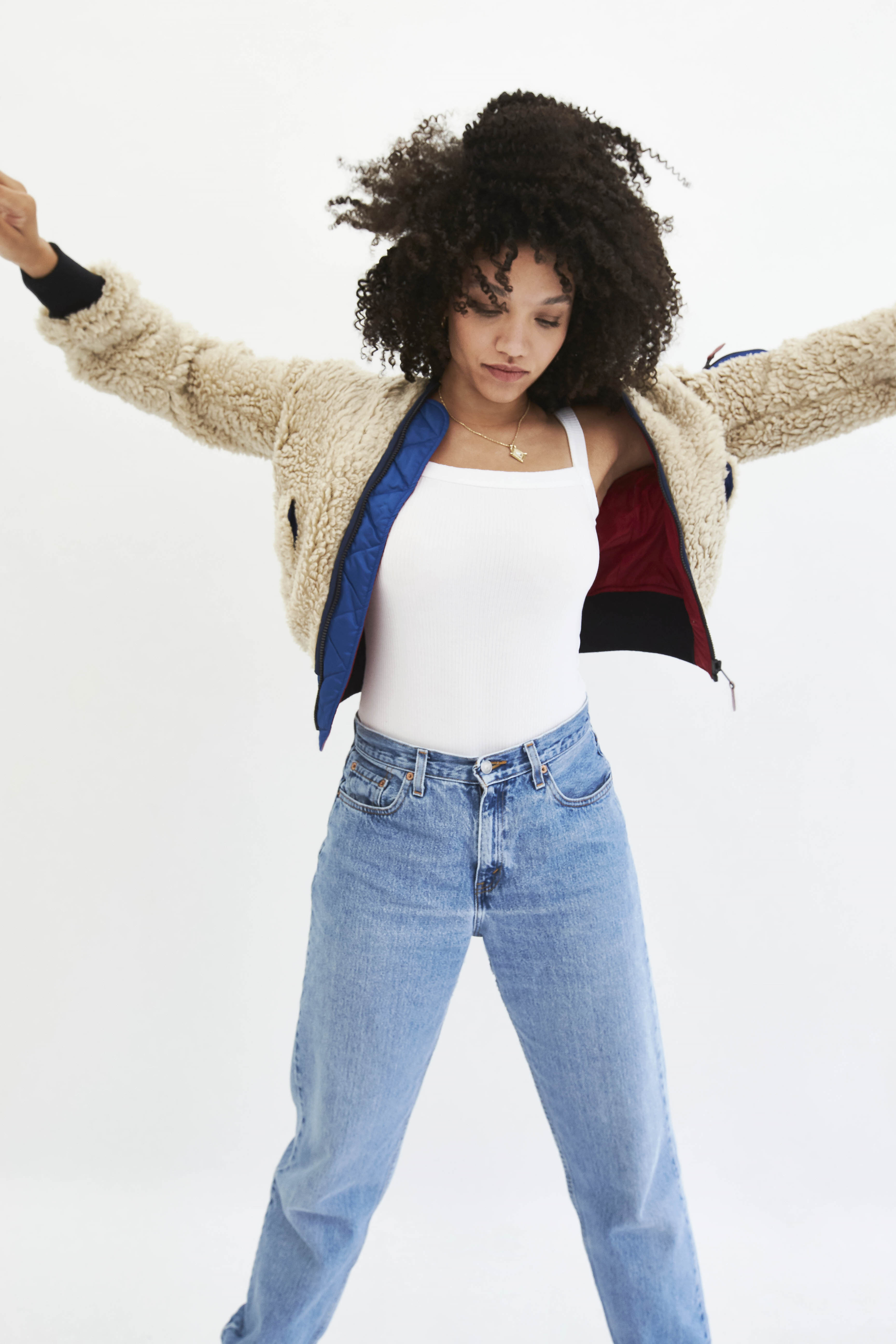 Woman with curly hair wearing a light jacket, a white tank top, and blue jeans in a full shot against a white backdrop.