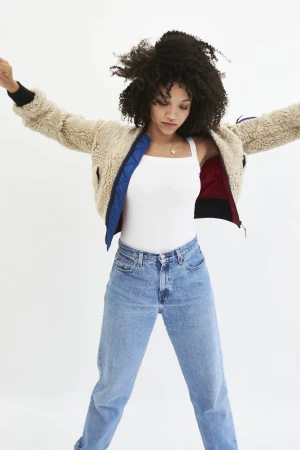 Woman with curly hair wearing a light jacket, a white tank top, and blue jeans in a full shot against a white backdrop.