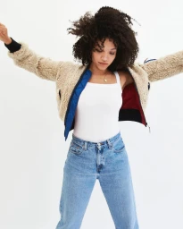 Woman with curly dark hair wearing a beige sherpa jacket, white tank top, and blue jeans against a plain white background.