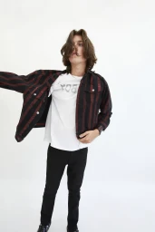 A young male model with long dark hair wearing a striped jacket, white t-shirt, and black jeans against a seamless white background.