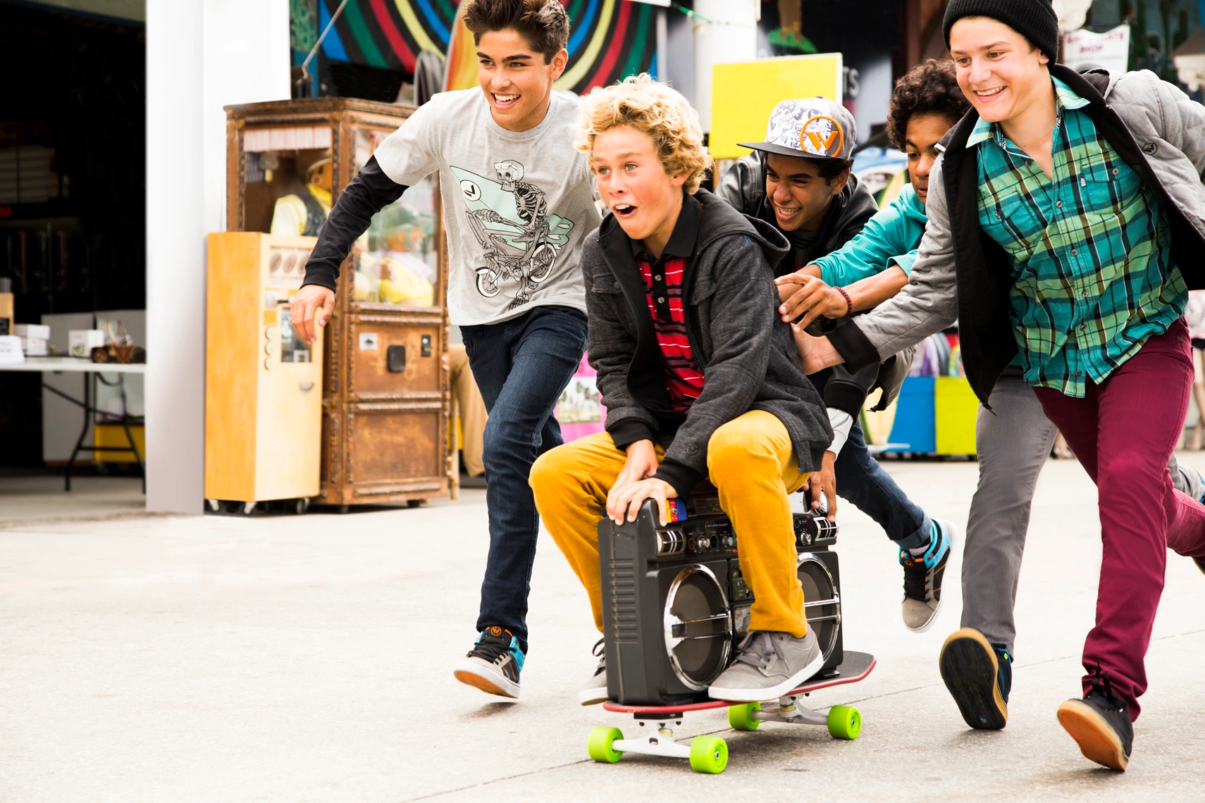 Group of teenage boys laughing and running outdoors while pushing a friend riding a skateboard loaded with a boombox.