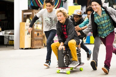 Group of teenage boys laughing and running outdoors while pushing a friend riding a skateboard loaded with a boombox.