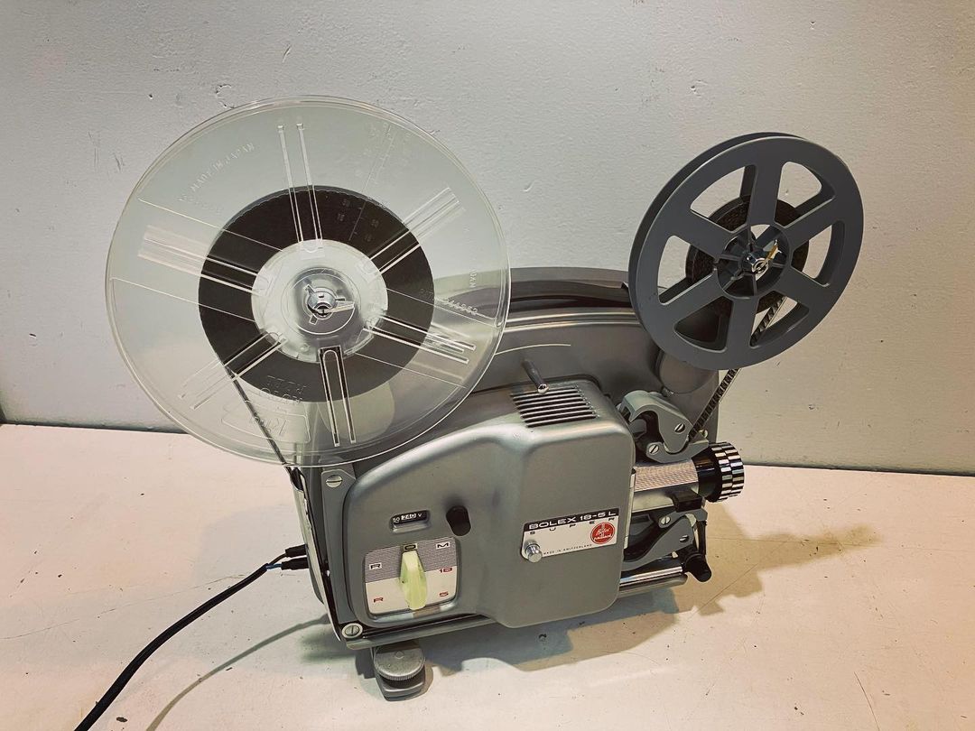 Vintage 8mm film projector displaying reels and film, standing on a table against a plain wall. A nostalgic piece of technology.