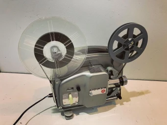 Vintage grey Bolex film projector with two reels resting on a plain white surface against a light grey wall.