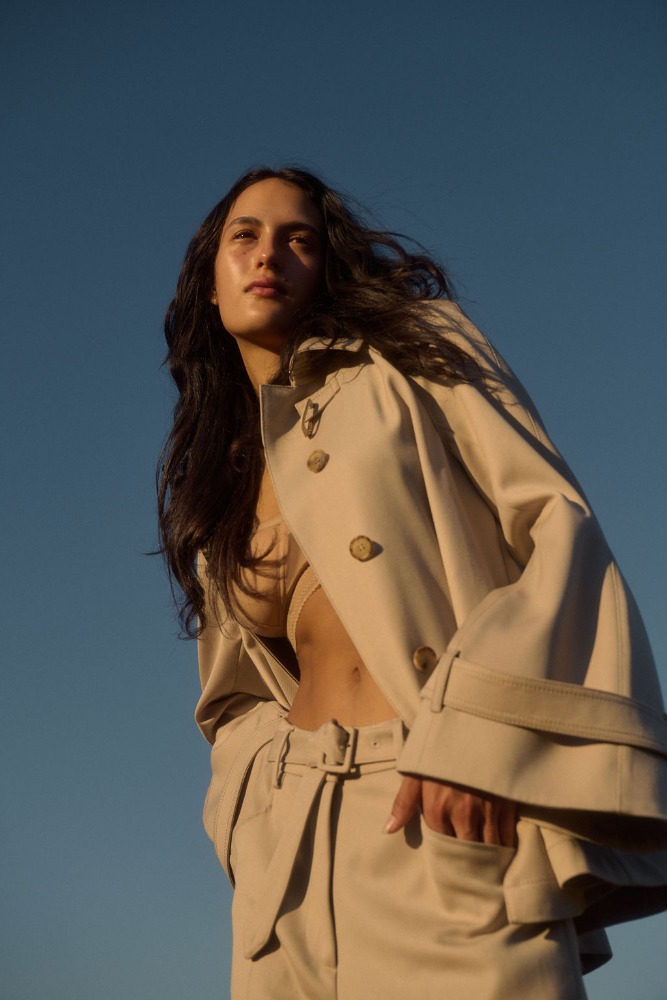Low angle shot of a stylish brunette woman in a beige trench coat and trousers against a clear blue sky, looking up and to the left.