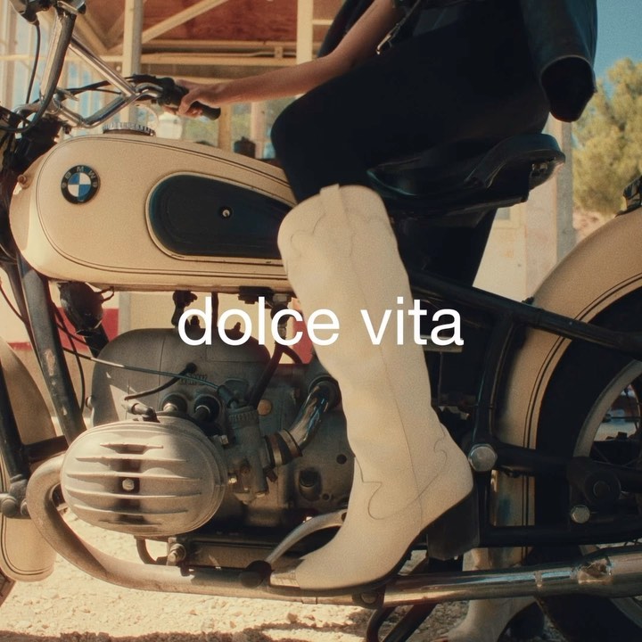 A woman in a vintage motorcycle with white boots, featuring the text 'dolce vita.' The motorcycle is light-colored, with natural outdoor light.