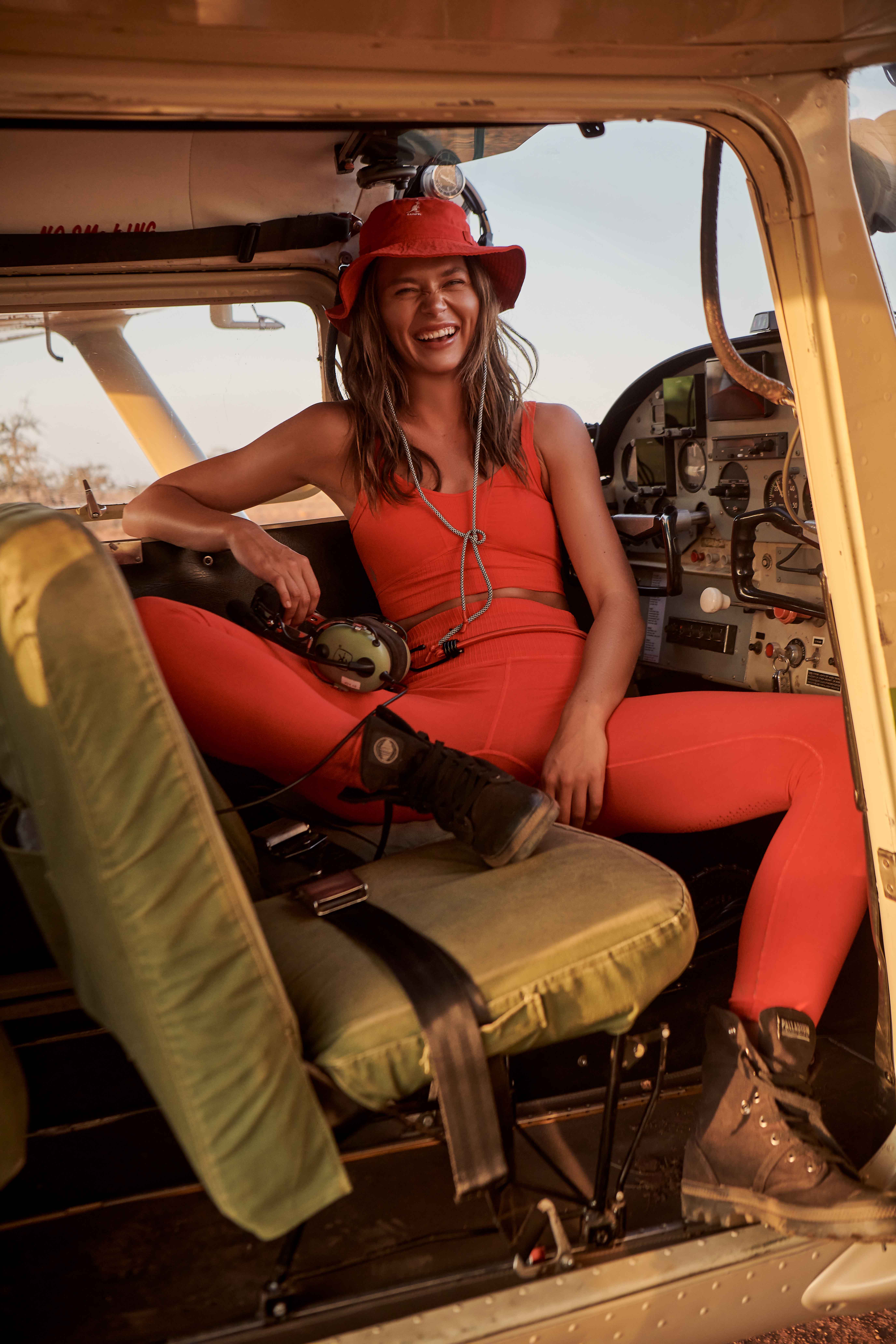 Smiling woman in orange activewear sitting inside a plane, wearing a hat and boots, near an open cockpit in a rural setting.