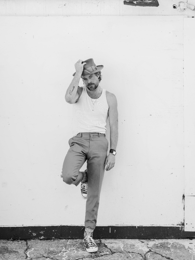 Monochrome portrait of a bearded man in a tank top and hat leaning against a white wall, wearing trousers and patterned sneakers.