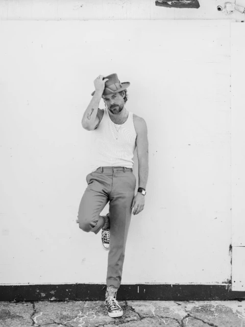 Monochrome portrait of a bearded man in a tank top and hat leaning against a white wall, wearing trousers and patterned sneakers.