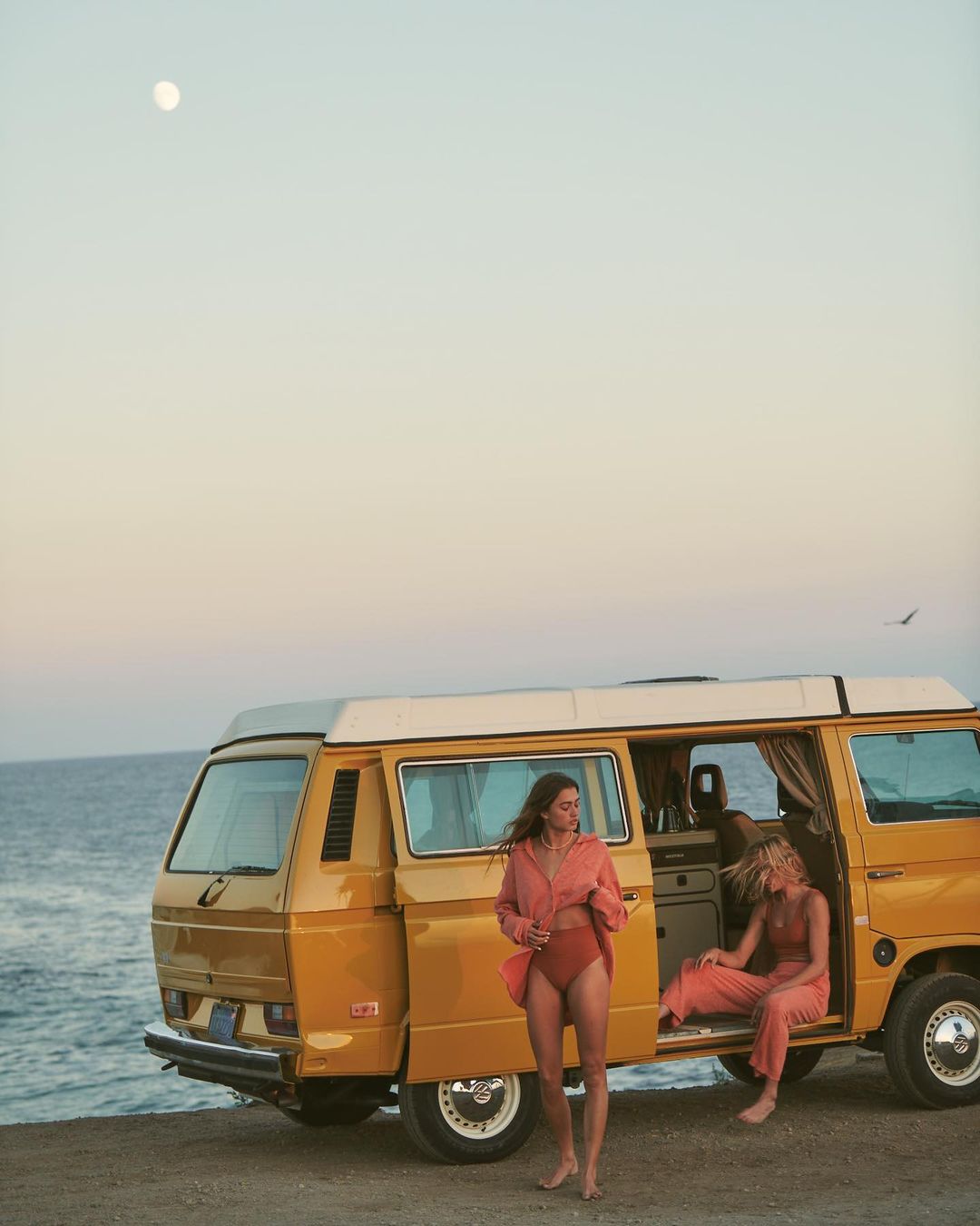 Two women in swimwear and casual shirts pose near a yellow camper van on a beach under a bright sky, bathed in golden hour light.