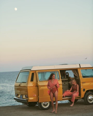 Two women in swimwear and casual shirts pose near a yellow camper van on a beach under a bright sky, bathed in golden hour light.