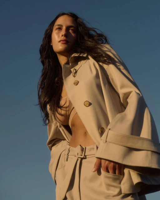 A woman poses in a beige coat and matching pants against a clear blue sky, looking up towards the light, showcasing fashion and beauty.