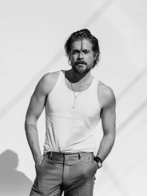 Black and white portrait of a man in a white tank top and grey pants posing in front of a white wall with light and shadows.
