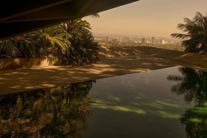 An architectural view overlooking a reflective swimming pool, palm trees, and a distant city skyline under an orange-toned sky.