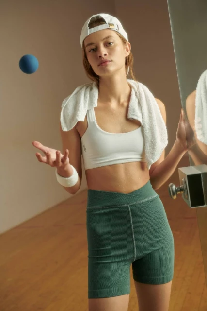 Woman in sportswear with a towel around her neck, wearing a baseball cap, holds a blue ball in a studio with mirrored wall.