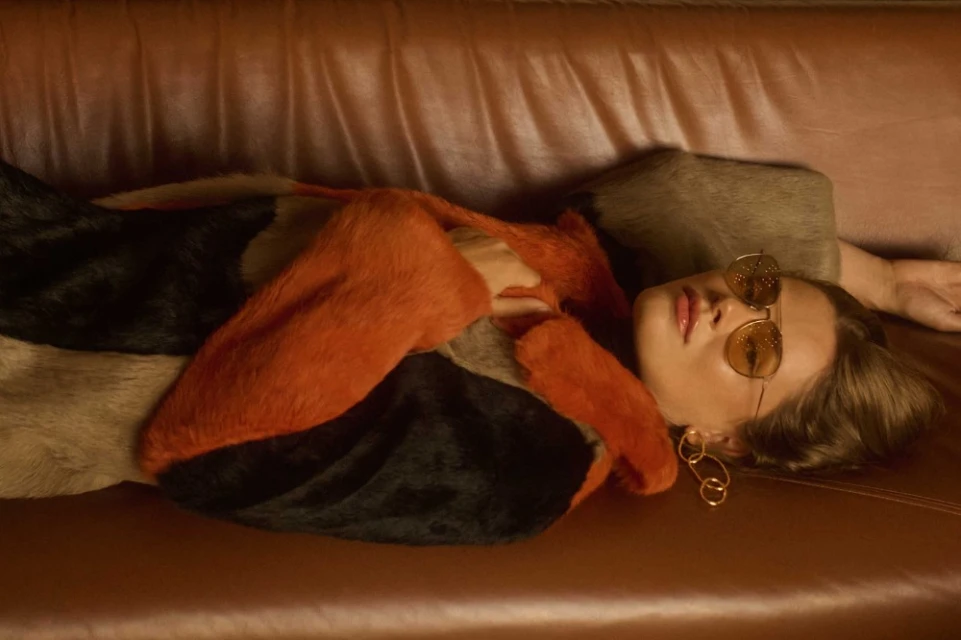A fair-skinned adult female lies on a peach-colored satin couch wearing sunglasses, orange top and multicolor coat with gold earrings.