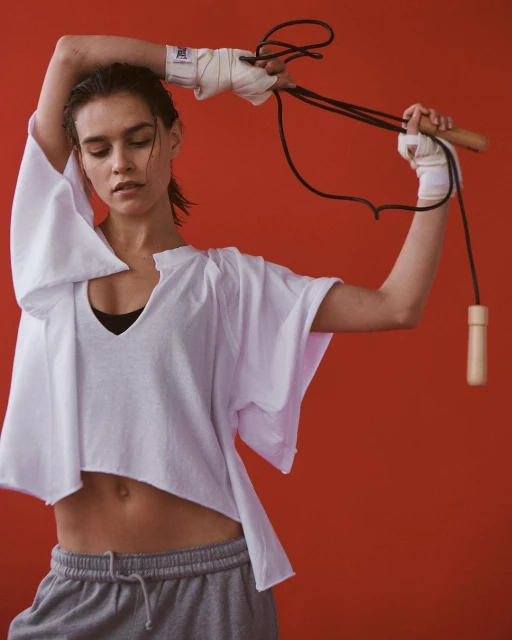An adult female with wet hair and hand wraps holds a jump rope in front of a solid red background. She is wearing a white t-shirt and grey sweatpants.