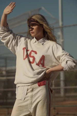 Blonde woman wearing white USA sweatshirt raising her hand to block the sun against an outdoor chain-link fence.