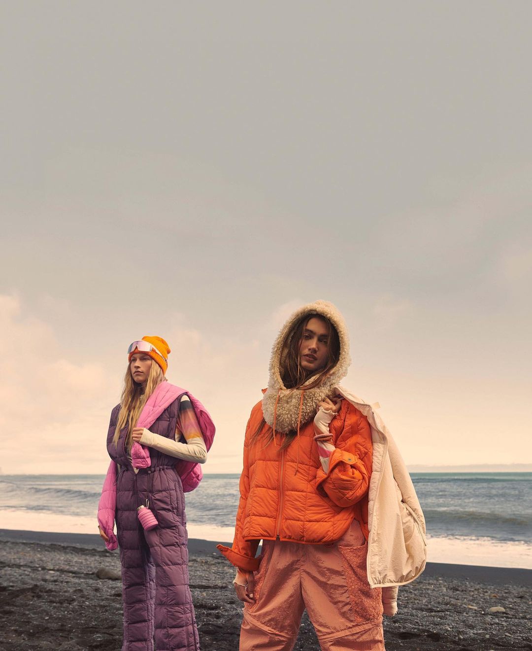 Two women in puffer jackets and snowsuits pose on a black sand beach with ocean and a cloudy sky in the background; fashion outdoor shoot.