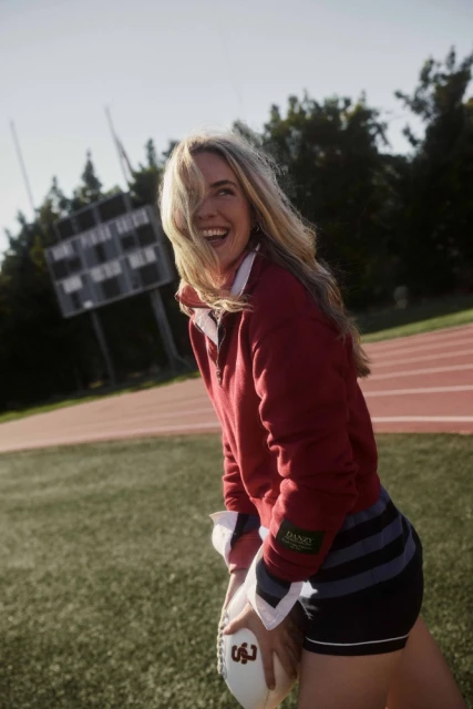 A smiling adult female wearing a red long-sleeve sports shirt and holding a football stands on a sports field, on a sunny day.