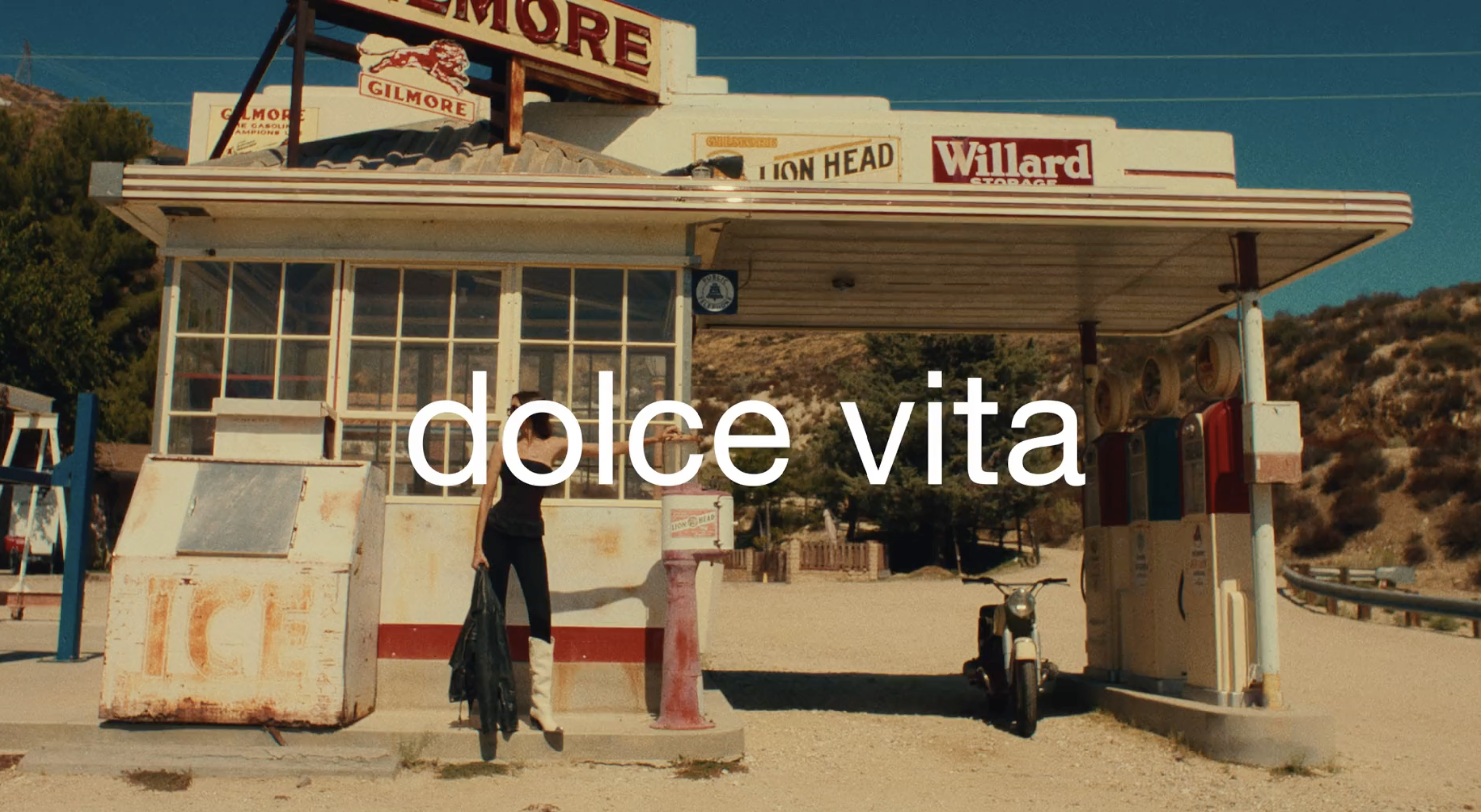 A woman stands near an old-fashioned gas station in a desert-like setting, holding a jacket with a vintage motorcycle nearby.
