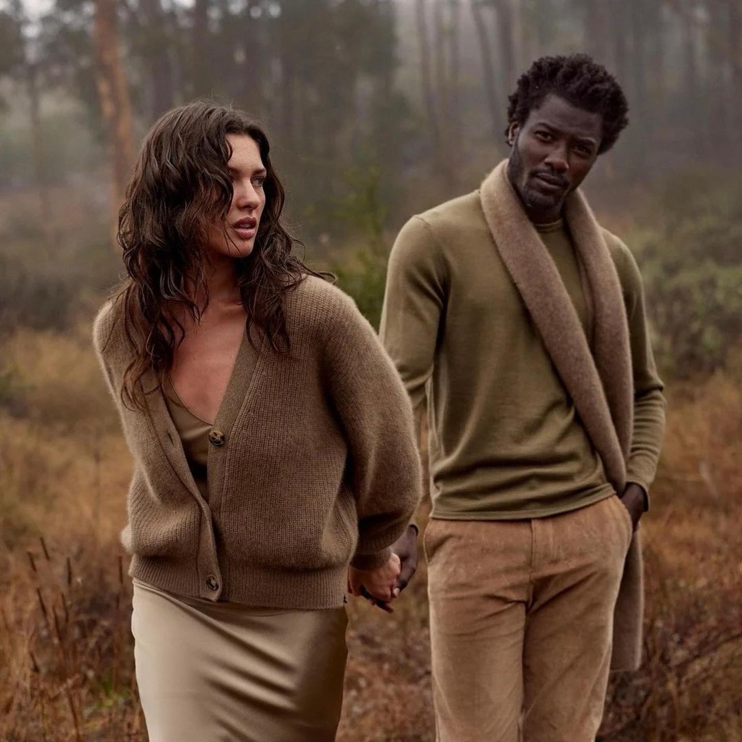 A couple stands in a misty forest wearing olive-green sweaters and neutral-toned pants and skirts, holding hands and looking off-camera.