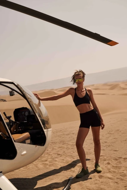 Woman in activewear poses near a white helicopter in a desert landscape under a clear sky, wearing sunglasses and athletic shoes.