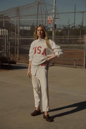 Woman in an oversized USA sweatshirt and light trousers standing on an outdoor asphalt court by a chain-link fence