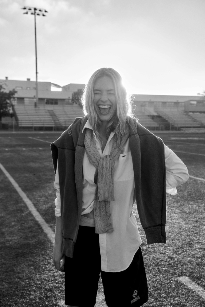 Black and white photo of an adult female laughing outdoors, wearing a sweater, button-down shirt and shorts on a grass field.