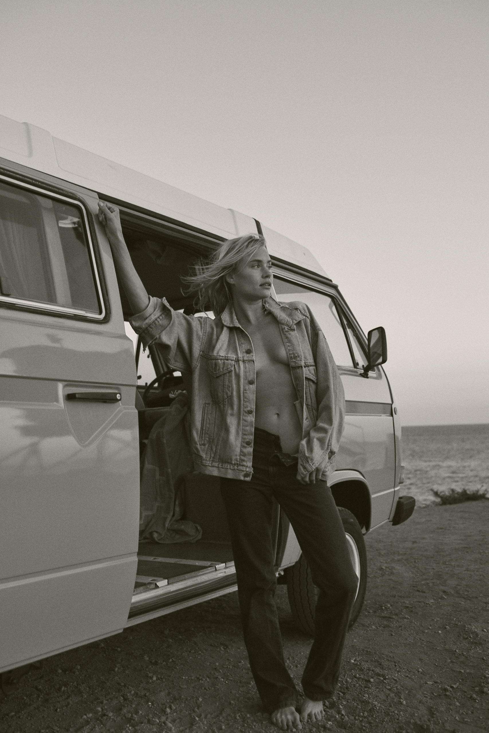 Blonde woman in denim jacket stands barefoot by van at beach. Sea and sky in background in vintage monochrome travel aesthetic.
