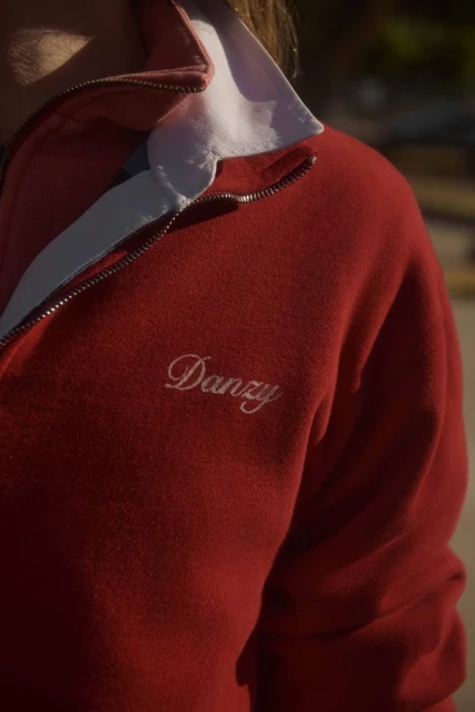 Close-up of a red quarter-zip sweater with a white collar, featuring 'Danzy' embroidered on the chest, captured in soft outdoor lighting.