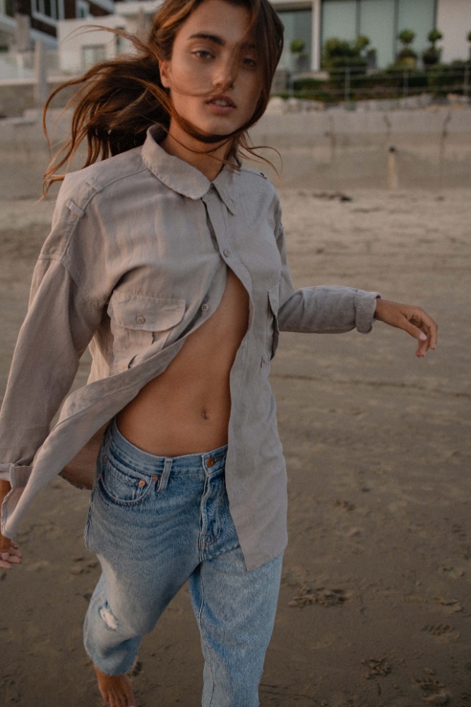 Woman walking on a sandy beach wearing a partially unbuttoned light grey shirt and blue jeans, photographed by Lindsey Childs.