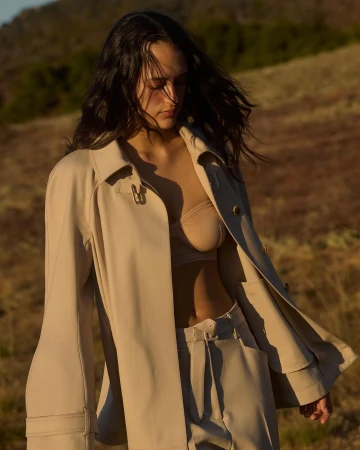 Woman in a beige trench coat and matching trousers posing outdoors in a dry grassy field during golden hour by photographer Lindsey Childs