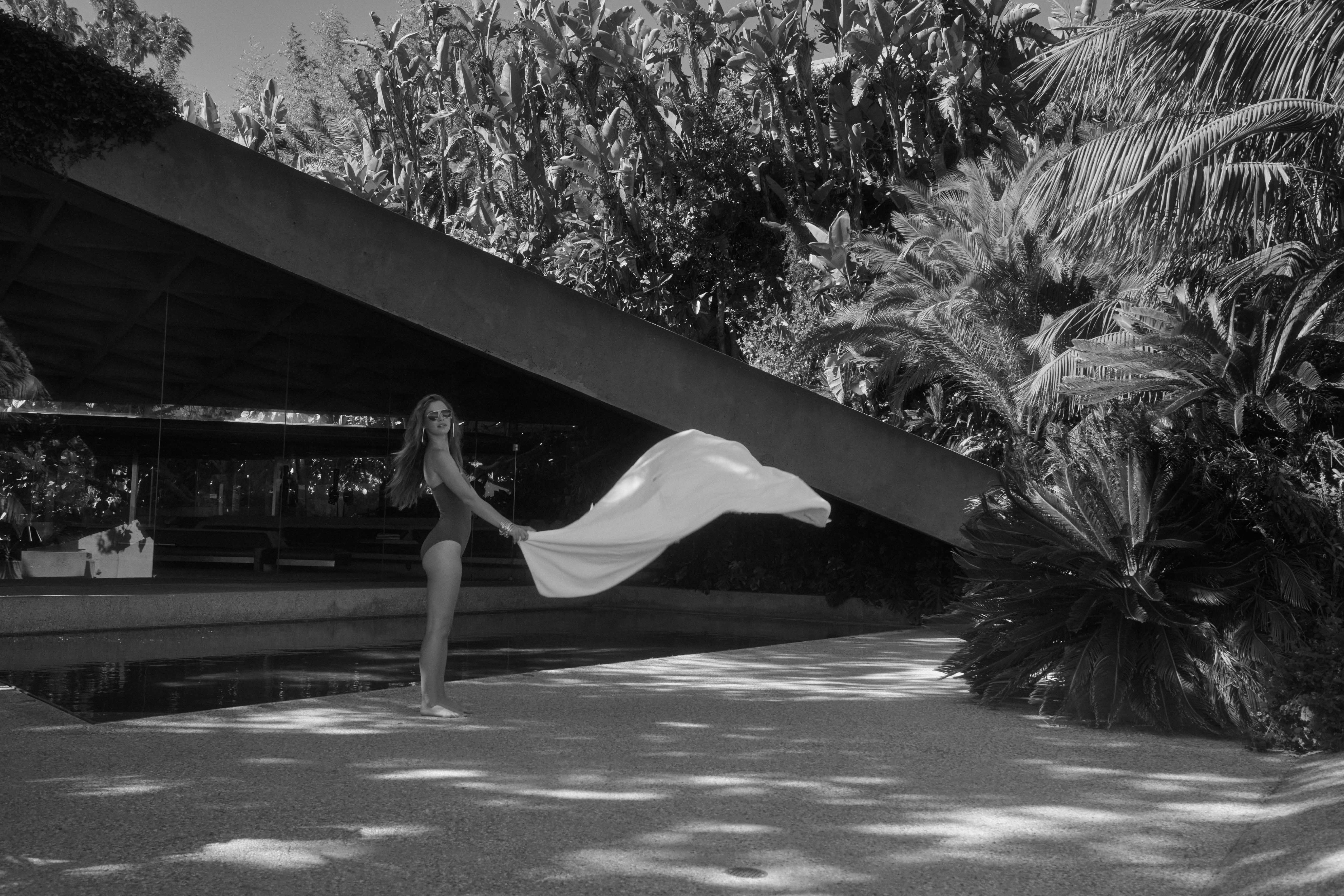 Monochrome shot of a woman in a swimsuit holding a white fabric, standing near a modern house and lush tropical vegetation.