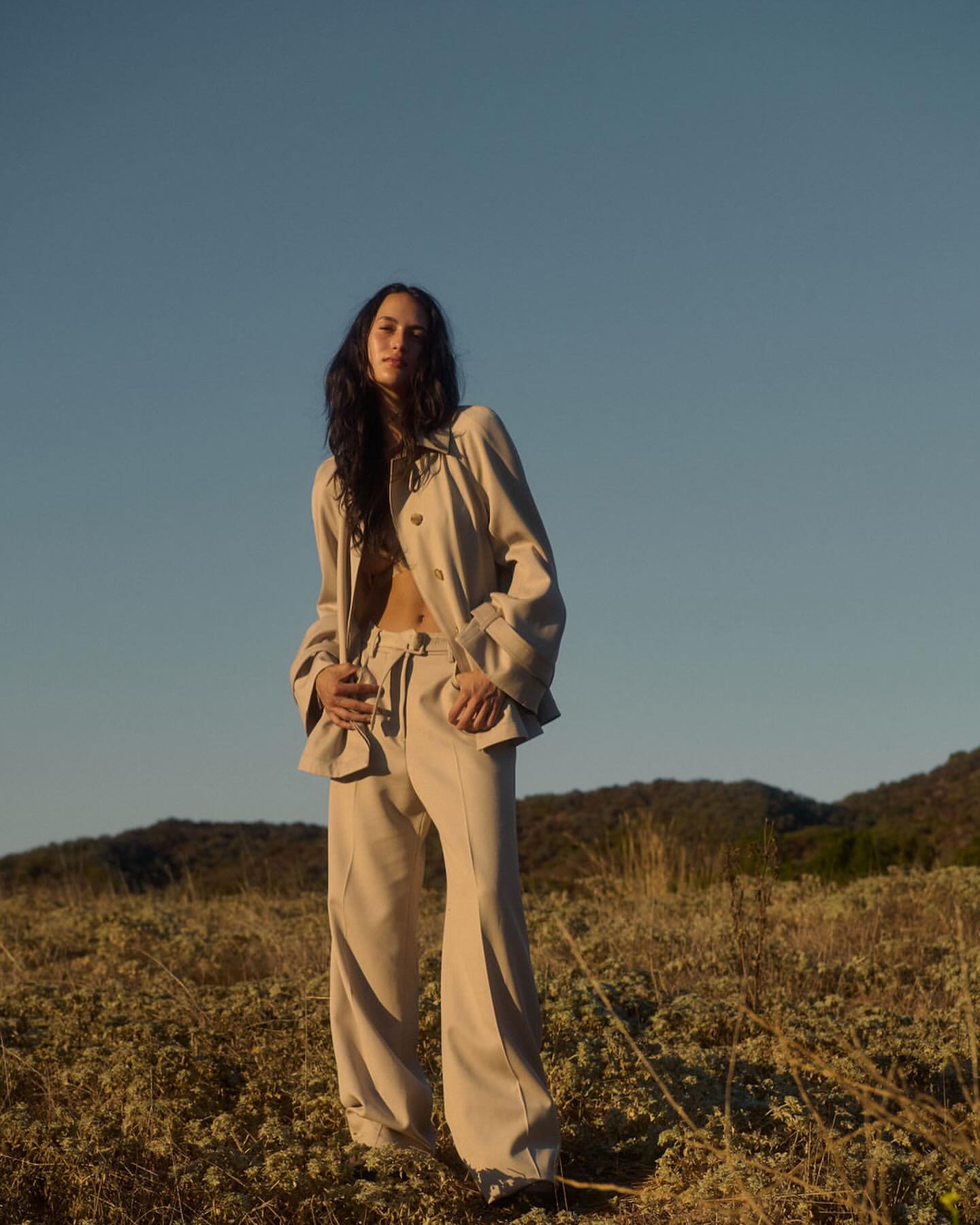 Woman in a beige button-down shirt and trousers standing in a grassy field under a clear blue sky