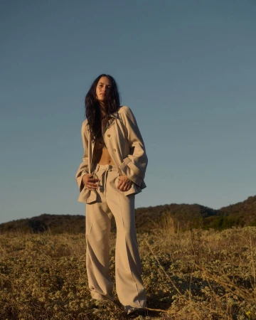 Woman in a beige button-down shirt and trousers standing in a grassy field under a clear blue sky