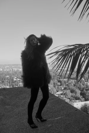 Monochrome portrait of a woman wearing a fluffy textured coat and high heels on a rooftop overlooking a city.