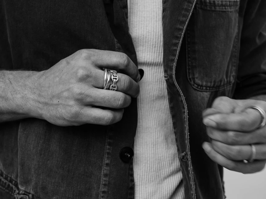 Close up of a male adult adjusting his denim jacket over a ribbed shirt, with stacked rings visible on his fingers.