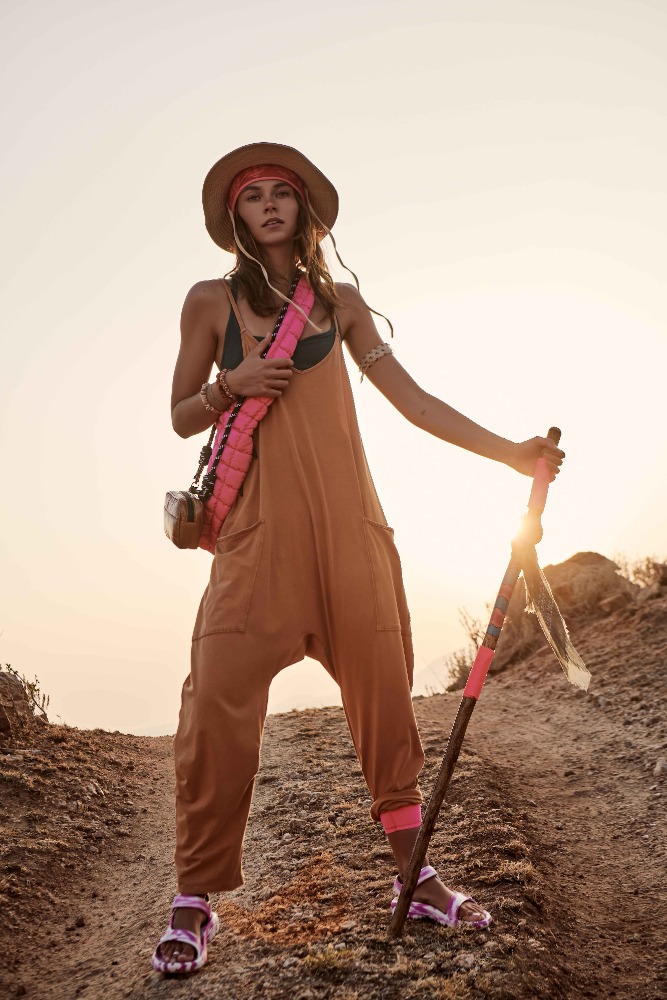 A woman with a walking stick stands on a dirt path wearing a hat, tan jumpsuit, and pink sandals, posing in an outdoor setting.