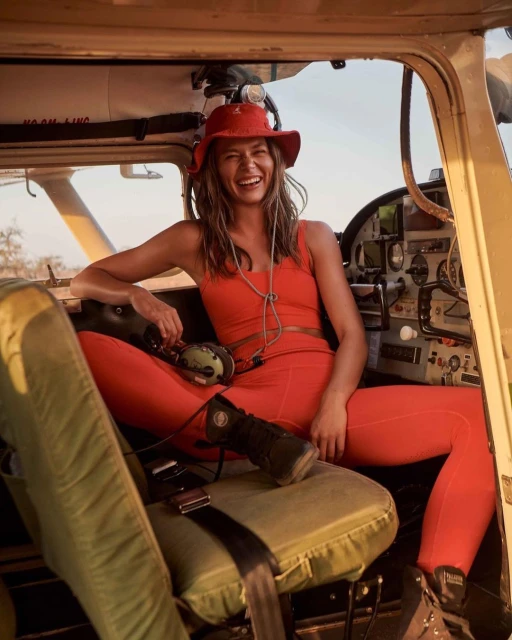 Smiling woman in red outfit and hat sits in airplane cockpit with a headset and olive-green seat and equipment in warm light.