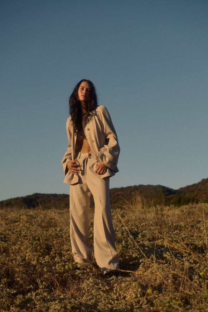 Woman in linen suit posing in field of wildflowers against blue sky in outdoor setting. Fashion editorial, lifestyle photography.
