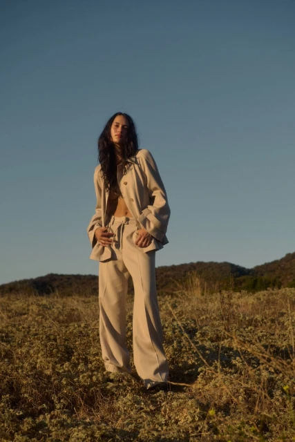 Woman in linen suit posing in field of wildflowers against blue sky in outdoor setting. Fashion editorial, lifestyle photography.