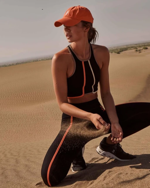 Woman crouching on sand dune wearing athletic top, leggings, baseball cap, and sneakers in an outdoor athletic fashion lifestyle shot.