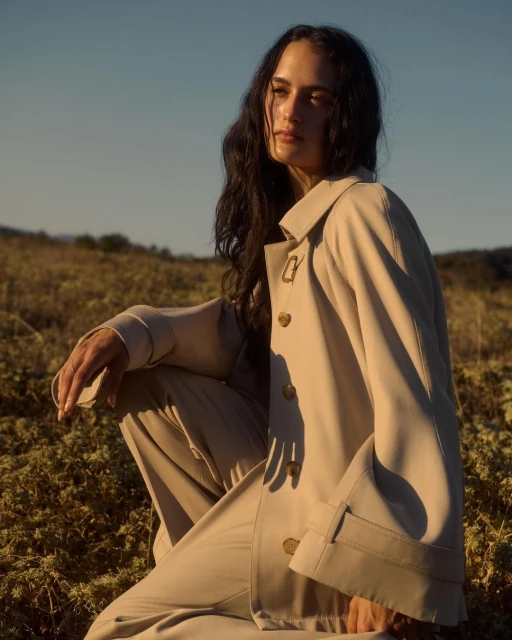 Young woman in a beige coat sits in a field under the sunlight, her dark hair cascading as she looks off to the side in a portrait
