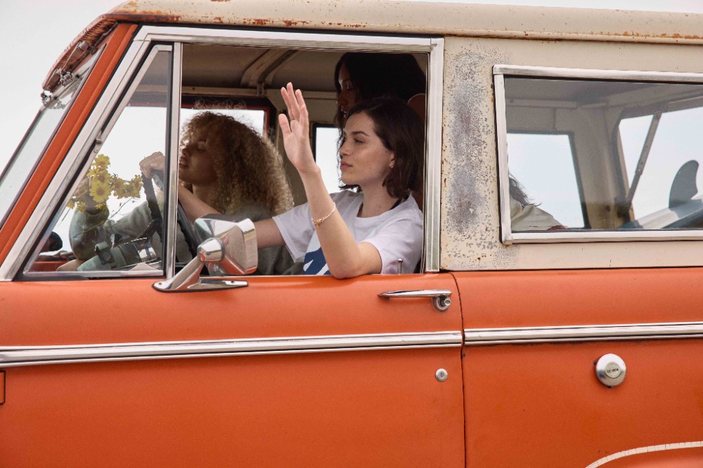 Three women inside an orange vintage SUV, one waving from the driver's seat, another with curly hair behind the steering wheel, and a surfboard.