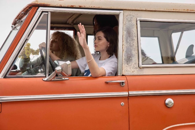 Three women inside an orange vintage SUV, one waving from the driver's seat, another with curly hair behind the steering wheel, and a surfboard.