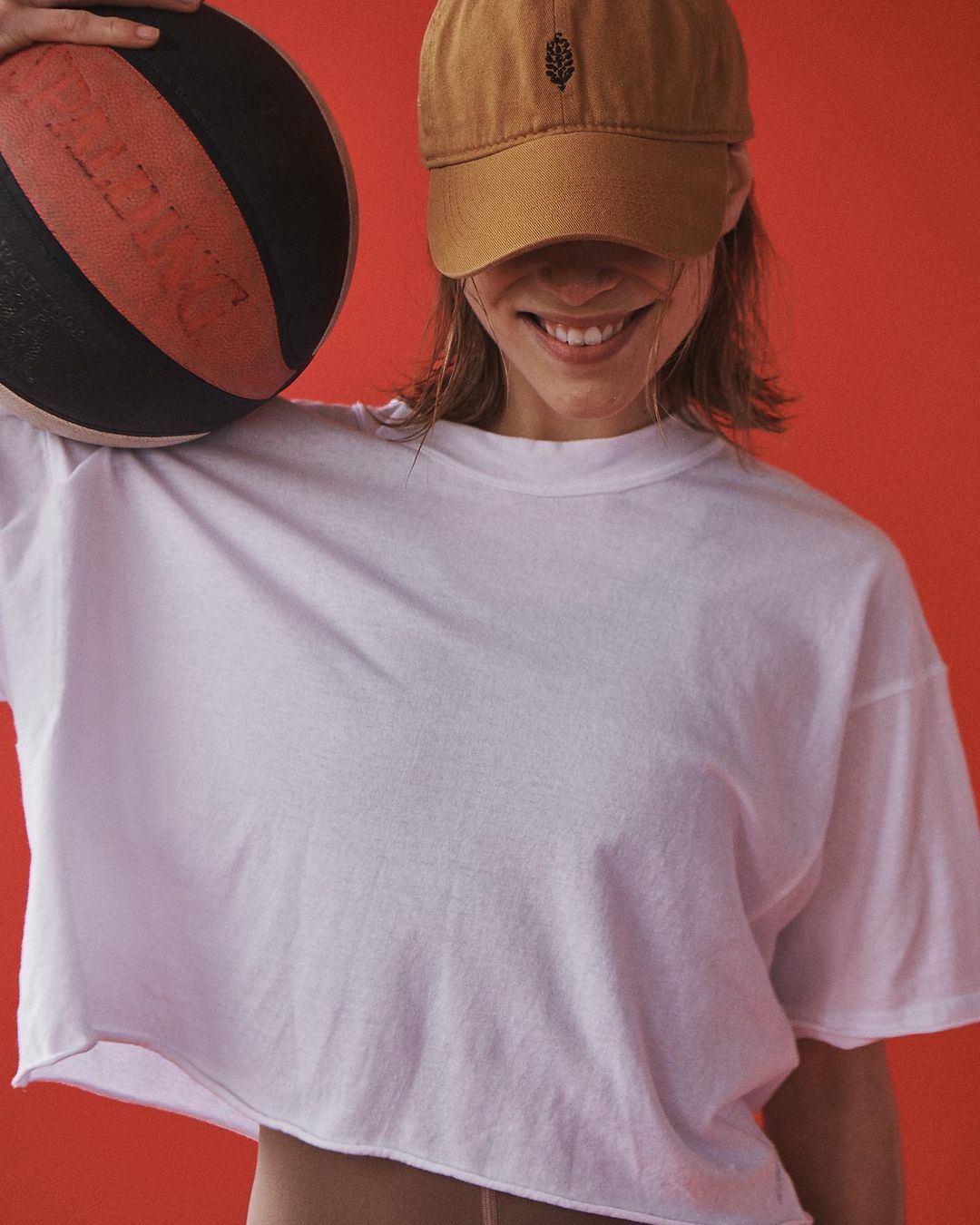 Smiling woman wearing a brown cap and white t-shirt holding a basketball against a solid red background.