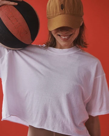 Smiling woman wearing a brown cap and white t-shirt holding a basketball against a solid red background.